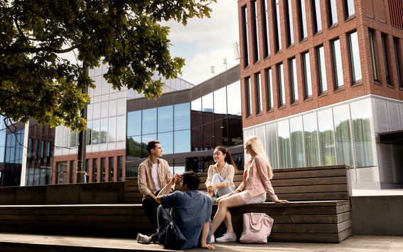 Four students sitting outside the Aalto University Väre building on wooden benches, green leaves from a tree is seen in the foreground.