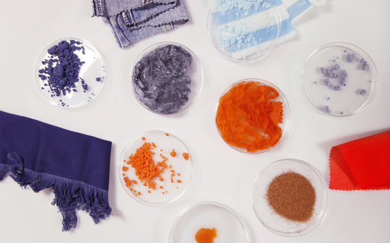 Laboratoriomaljoissa on erilaatuisia ja erivärisiä tekstiilikuituja ja niiden vierssä pöydällä kangastilkkuja, muun muassa farkkukangasta.