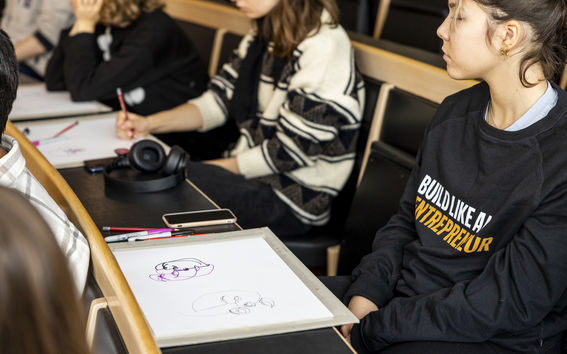 A person wearing a shirt that says "build like an entrepreneur" sitting in class, in front of a drawing