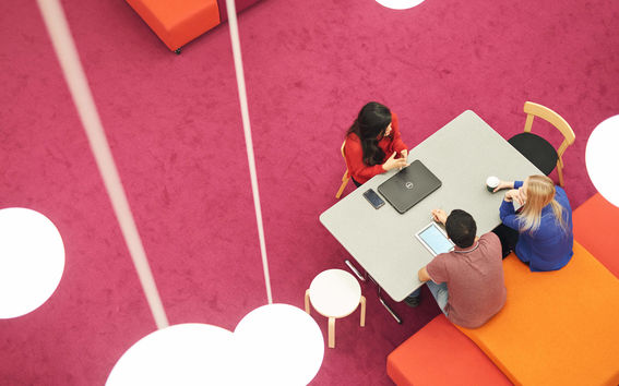 Three students having a discussion around a table at the Learning Centre at Aalto. Around the edges, there are also colourful furniture and lamps. Photo: Aalto University / Unto Rautio.