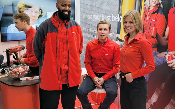 Two people standing and one sitting on a fair stand and all are wearing red shirts. A huge wall with picture of a woman with red shirt and Hilti logo on the background.