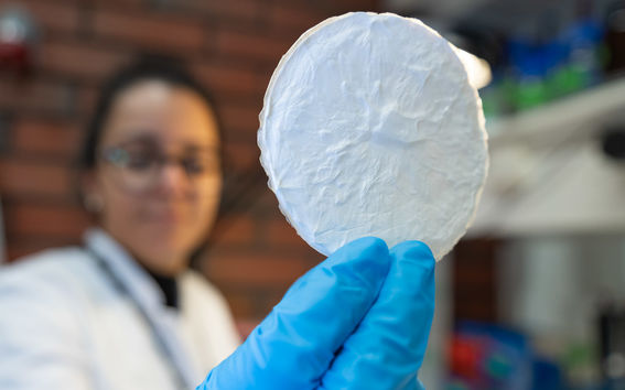 Colour photo of a blurry person in a white lab coat holding an in-focus white paper-like wafer in their blue gloved hand