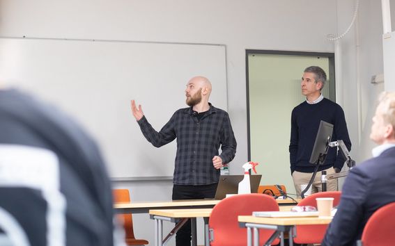 A researcher in dark clothing presenting his work in front of a classroom, gesturing towards the whiteboard and talking. 