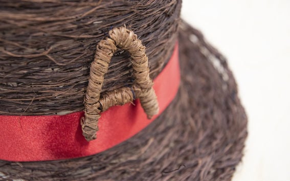 Color photo of a hat made of fine bark strips featuring a red hatband and a fiber-constructed letter "A"