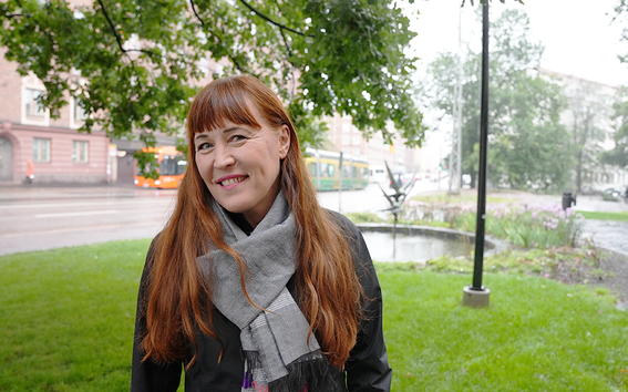 Red-haired woman standing outside of Aalto University Töölö building in Helsinki, surrounded by trees and grass. A bus and tram on the background.