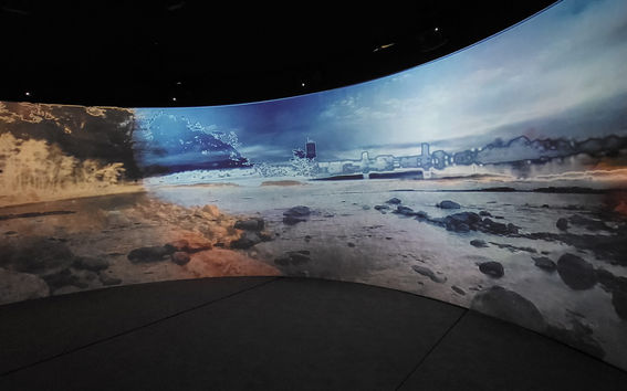 A video being played on a large, wrap-around screen. The video has a rocky coastline in the foreground and a blurry city skyline in the background, with water in between. The sky is bright, and the image has been distorted to enhance the edges of the coastline and a copse of trees on the left.