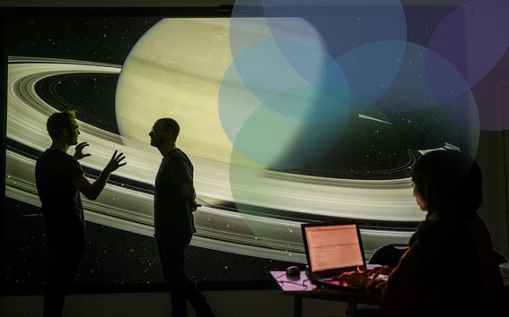 two people discussing in front of a space illustration, while another person takes notes on a laptop