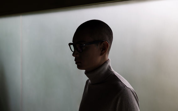 A man wearing glasses with heavy shadows covering his face stands in front of a grey wall.