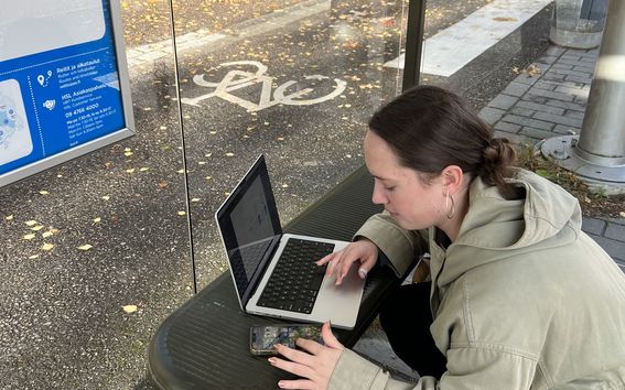 A woman is working on her phone and laptop at a bus stop