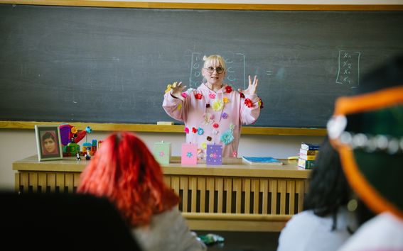Leikkisä matematiikan professori Pauliina Ilmonen seisoo opiskelijoiden edessä luokassa taustanaan liitutaulu. 