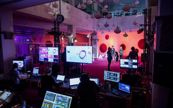 Laajassa kuvassa studiosta näkyy tila kokonaisuudessaan, etualalla tietokoneen näyttöjä sekä tekniikasta vastanneita ihmisiä kuulokkeet päässä, taustalla tilaisuuden juontajat ja muita taustahenkilöitä.