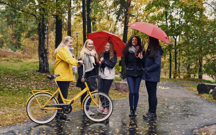 Five students standing in the rain. One has a bike and two have a red umbrella.