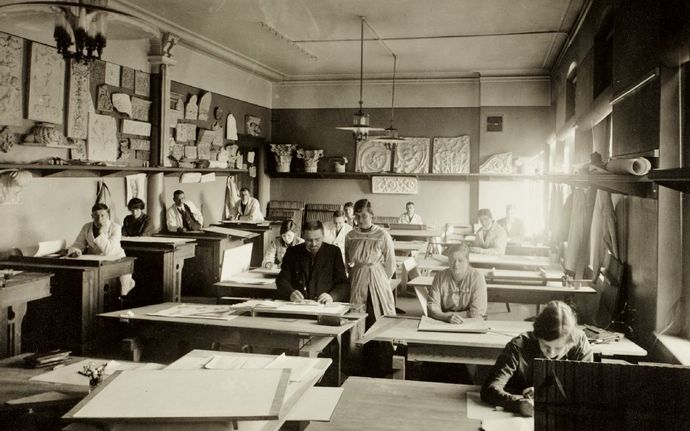 Mustavalkokuvassa vuodelta 1920 oppilaita ja opettaja istuvat veistokoulun oppitunnilla pöytien äärellä, yksi opiskelija seisoo opettajan vieressä.