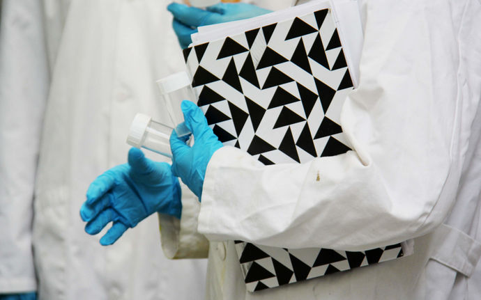 A person with a lab coat and protective gloves carrying test tubes and a black-and-white notebook. Their face is not pictured.
