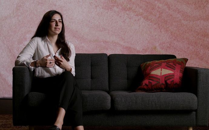 Natalia Villaman in long dark brown hair and a white shirt sitting on a dark grey sofa explaining and talking vividly with her hands towards the camera.
