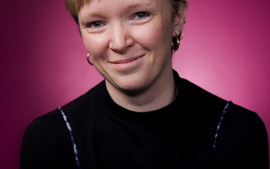 Portrait of Aalto student Henriette Friis who smiles in the picture and is wearing a black shirt and blue dress, sitting on a chair against a purple background.