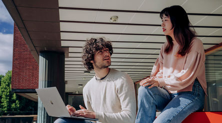Two summer school students discussing by a laptop outside a campus building.