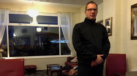 kristian in a black jacket and black glasses standing in a room with red sofas and chairs