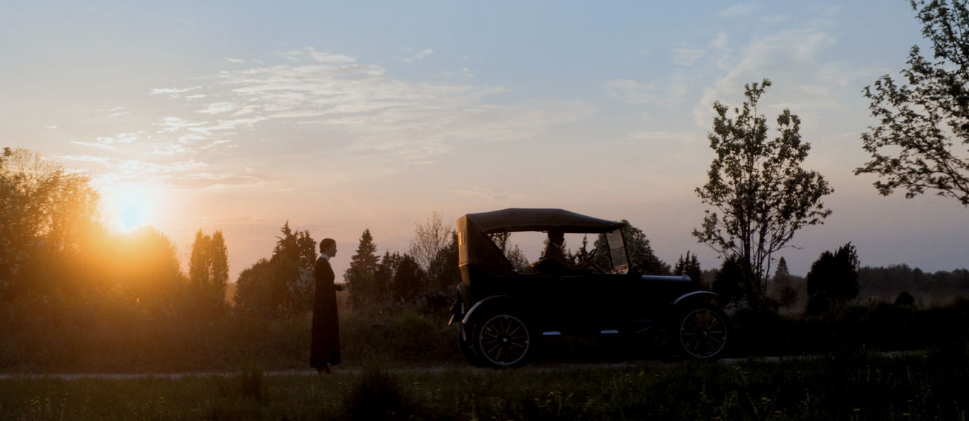 Helene-elokuvan kohtauksessa näkyy laskevan auringon maisemassa vanhanaikainen auto ja sen takana nainen seisomassa pää hieman painuksissa.