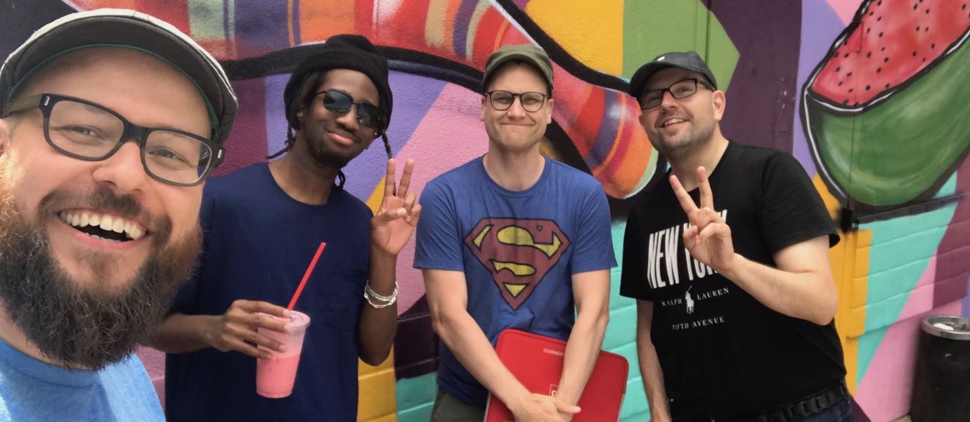 Four smiling men posing in front of a colorful graffiti wall. 