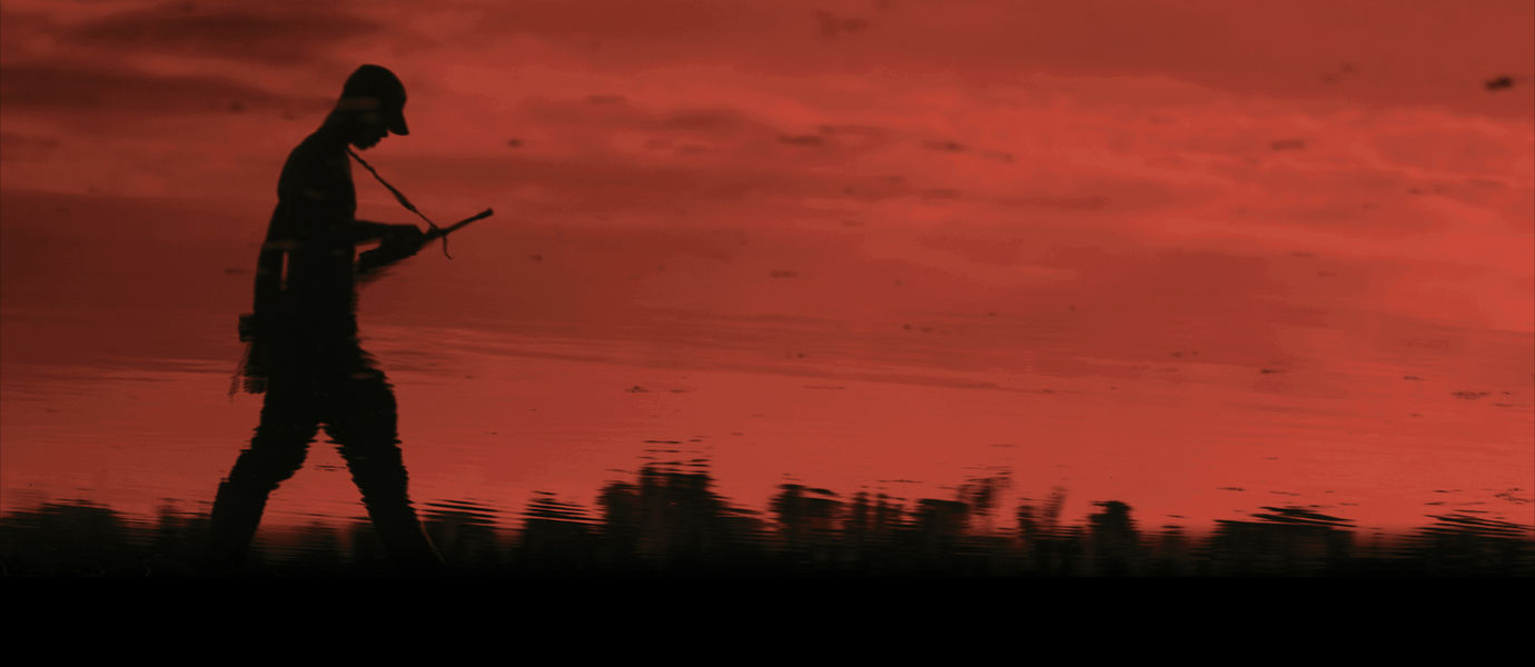 Tumma aseistautunut mieshahmo kävelee punaisen auringonlaskun maisemassa 
