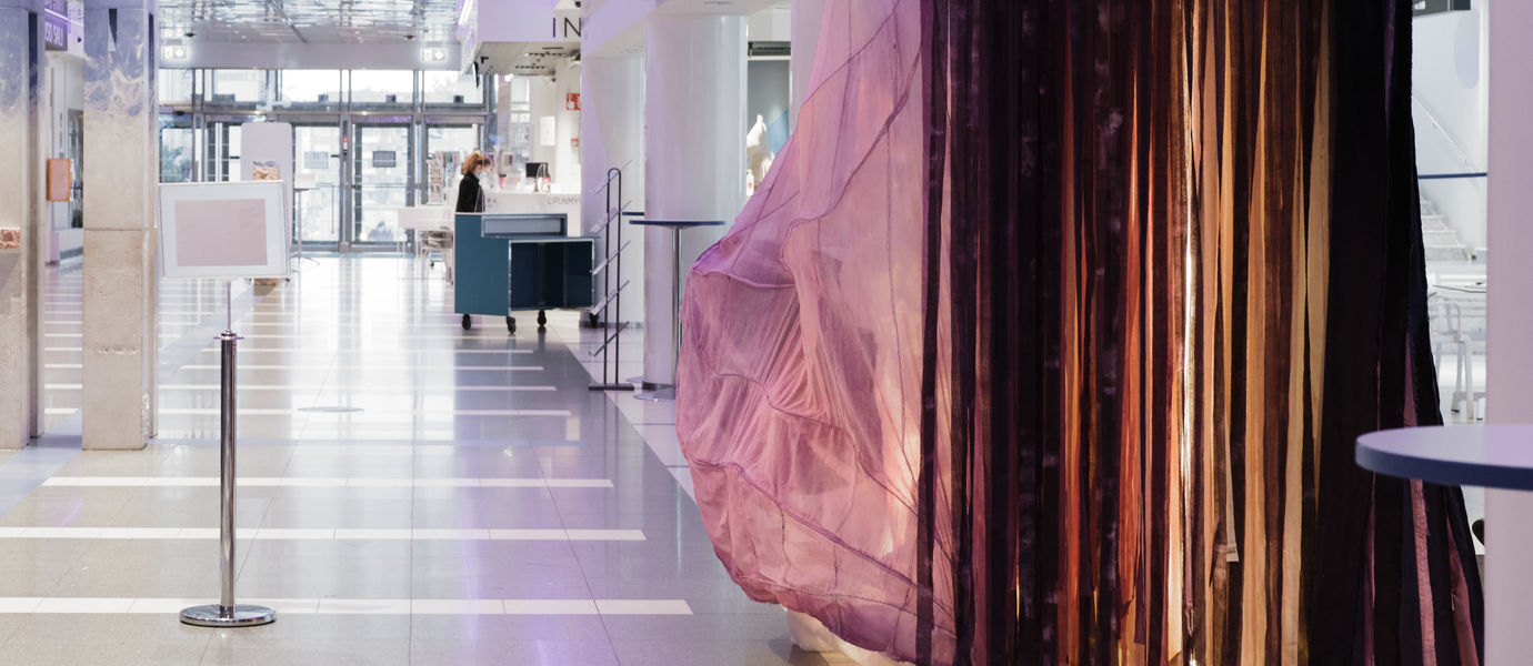 Kuvassa taideinstallaatio SOONI vaaleassa aulatilassa. Teoksen värikkäät tekstiilit liikkuvat teoksesta kantautuvan äänivirran mukana.