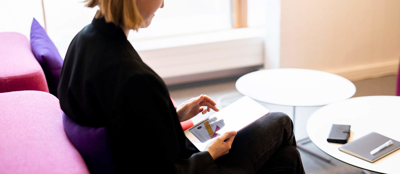 Toimistossa istuva nainen selaa tablettiaan, pöydällä esillä puhelin, muistikirja ja kynä.