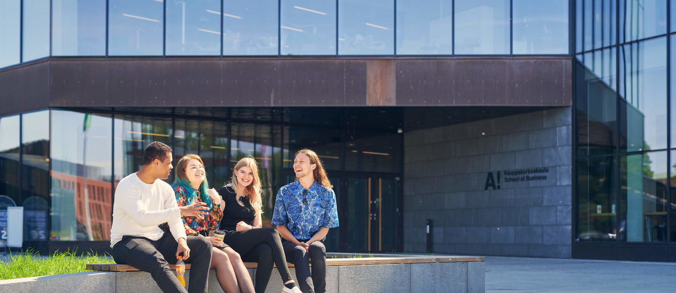 Four students sitting in on a wall outside the Aalto University School of Business building. They are all laughing and discussing vividly.