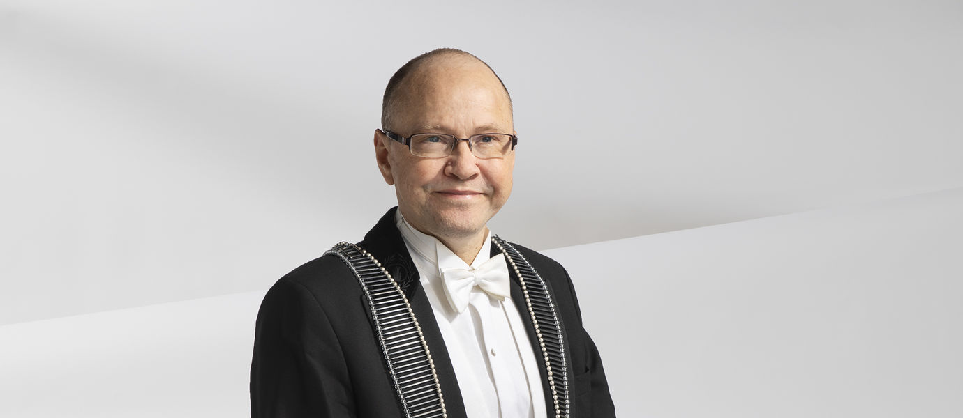 Janne Laine sits on a tall black chair, dressed in a tuxedo, wearing the Dean of School of Cheminal Engineering ceremonial link necklace, smiling. Background is white and slightly wavy.