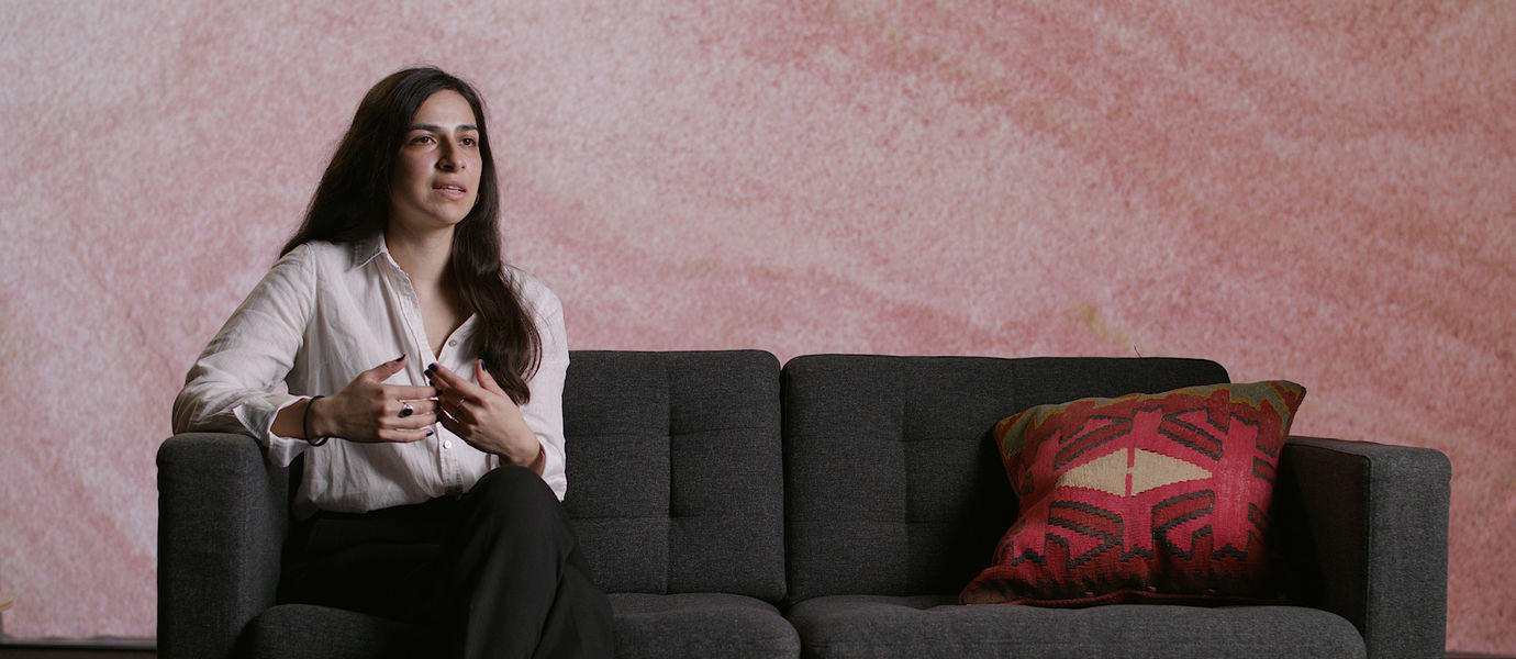 Natalia Villaman in long dark brown hair and a white shirt sitting on a dark grey sofa explaining and talking vividly with her hands towards the camera.