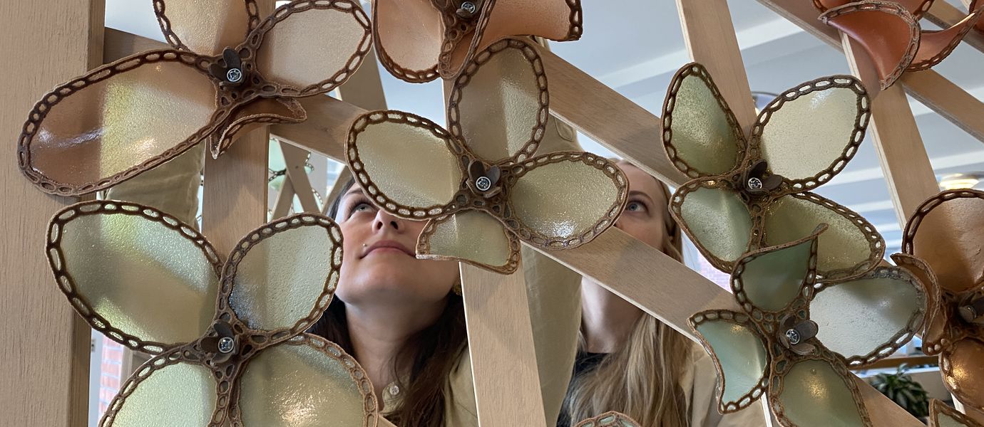 Two young people are looking upwards and smiling with mouths closed, behind an installation they designed. Art is made of wood and biopolymer flowers.