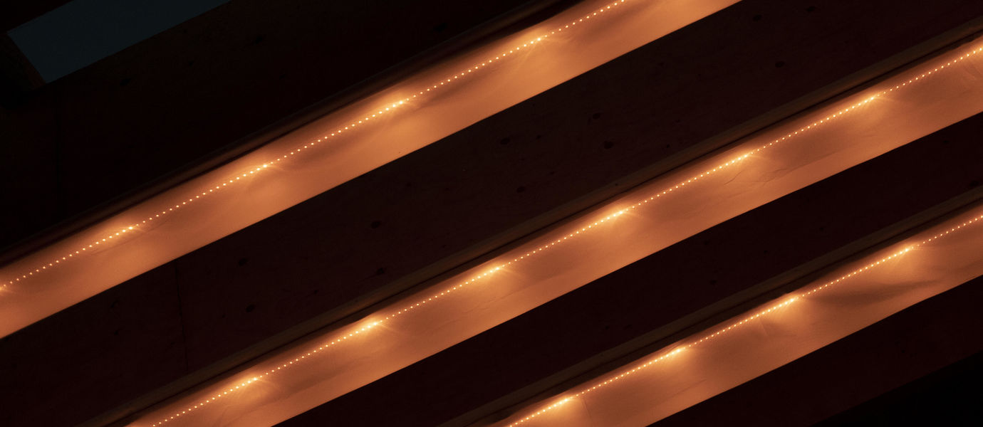 Three light fixtures lit up on the ceiling of a structure, looks like three beige stripes on a black background, going across from left down corner to right upper corner.