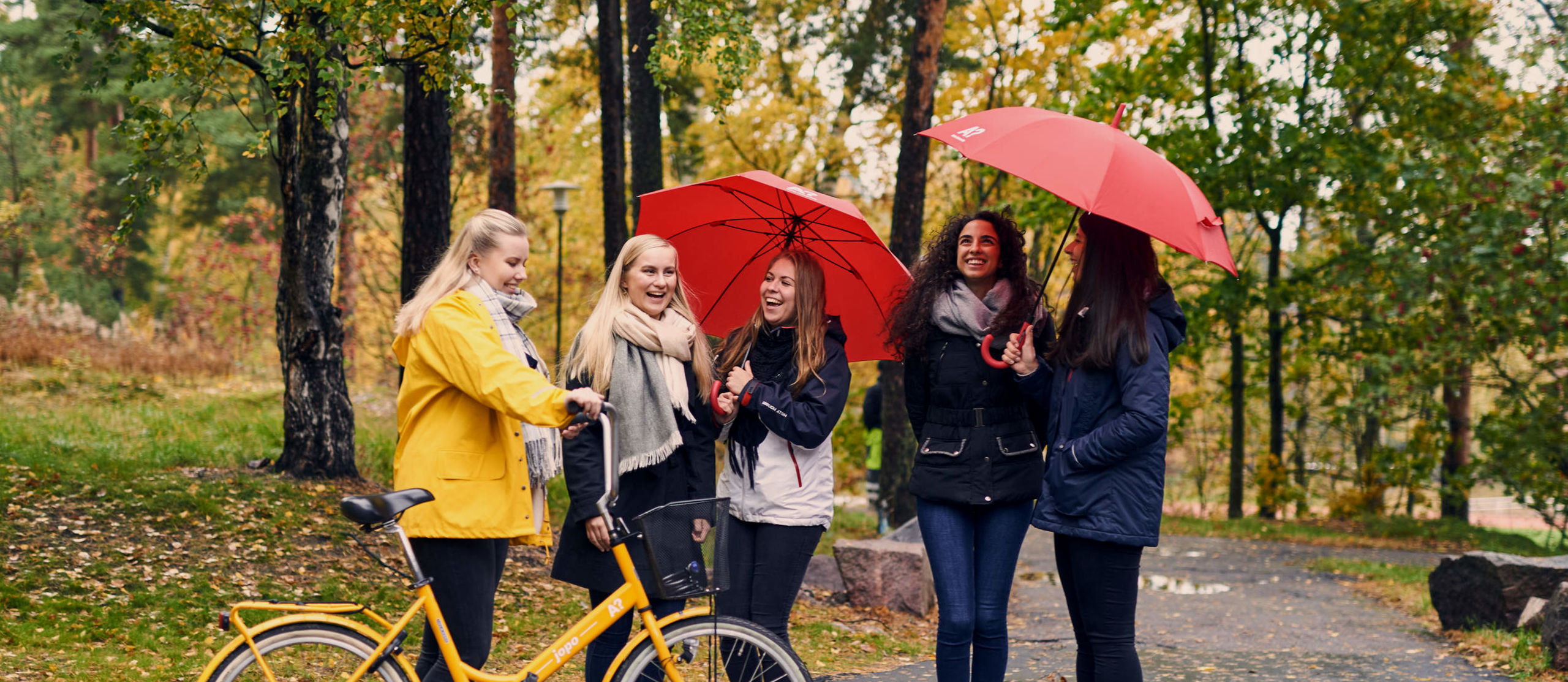 Five people with a yellow bike and red umbrellas on autumnal campus