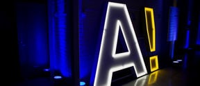 Human-sized lamp in the shape of the Aalto logo, capital A followed by an exclamation mark, stands in a dark space / photo by Aalto University, Lasse Lecklin