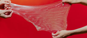 kuvassa kahdet kädet levittävät läpinäkyvää limaa punaista taustaa vasten