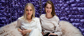 kaksi naista istuu vaalealla karvataljalla violeteista putkista koostuvan taustan edustalla ja katsoo kameraan