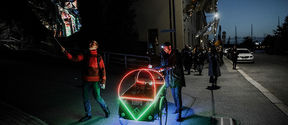 A photo of a performance where a person is pushing a two-wheeled cart in the street. The cart is lit with neon lights, around are people who are waving silver flags.