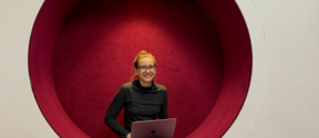 Svitlana Chaplinska sitting with her laptop and smiling towards the camera in a futuristic, round and red chair integrated to the wall at Aalto University campus.