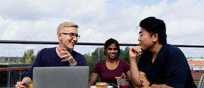 Three students eating a snack and discussing over a computer on a rooftop terrace. 