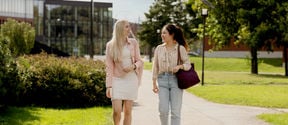 Two female students walking in a lush and green Aalto University campus, looking at each other while talking and walking.