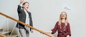 Marjo-Riitta Diehl ja Miisa Mink seisovat vaaleassa portaikossa rappusilla. Diehl on heittänyt ilmaan paperilennokin, joka leijailee Minkin kasvojen yläpuolella. Molemmat hymyilevät. 
