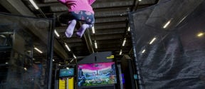 A young girl in mid-jump on a trampoline. She is facing away from the camera with her knees up and her arms extended. Facing the trampoline is a television showing a video game in which the young girl is in mid-jump above another character.