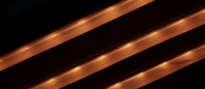 Three light fixtures lit up on the ceiling of a structure, looks like three beige stripes on a black background, going across from left down corner to right upper corner.