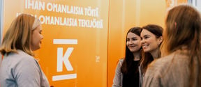 Four people standing infront of orange-colored walls with texts and K group logo. A bowl of candy.