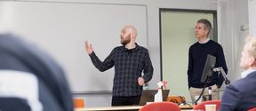 A researcher in dark clothing presenting his work in front of a classroom, gesturing towards the whiteboard and talking. 