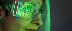 A closeup of a woman in green light wearing goggles that reflect colourful pixels 