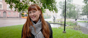 Red-haired woman standing outside of Aalto University Töölö building in Helsinki, surrounded by trees and grass. A bus and tram on the background.