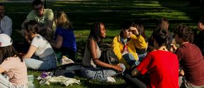 A group of undergradiate summer school students during a welcome event picnic on a summery day at Aalto University campus. The sun is shining and the group of students are sitting in on the grass in a park-like setting.