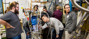 A group of seven researchers observe a complex piece of machinery in the center of the photo.