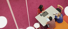 Three students photographed from above, students dressed in red and blue sitting at a table in a room with a pink floor